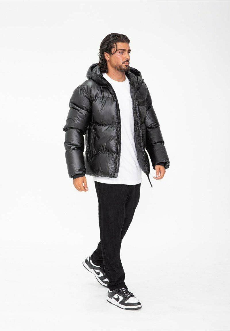 Doudoune noire avec une finition brillante, capuche et coutures bien visibles. Portée sur une chemise blanche, associée à un pantalon noir et des baskets noir et blanc.