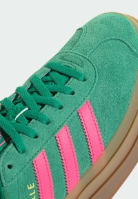 Gröna mockaskor med rosa zickzackränder, texturerad ovandel och matchande gröna snörningar. Har en tan gummisula.