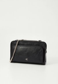 Sac en cuir noir Calvin Klein avec poche zippée à l'avant et bandoulière chaîne argentée sur fond blanc.