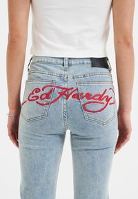 Lyseblå denim jeans med synlige baglommer. Funktioner stort rødt broderet "Ed Hardy"-tekst hen over ryggen. Klassisk fem-lomme design.