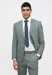 Gaiši pelēks uzvalks ar strukturētu dizainu, divām pogām un sānu kabatām. Apvienots ar baltu krekla un jūras zilas svītrainas kaklasaites. Pielāgota piegriezums.