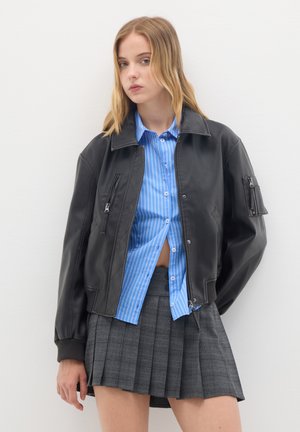Jeune femme aux longs cheveux blonds portant une veste en cuir noire, une chemise rayée bleue et une jupe plissée grise sur un fond blanc.