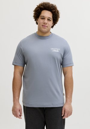 Άνδρας με σγουρά μαλλιά και φακίδες φοράει ανοιχτό μπλε μπλουζάκι "Originals Studio" και σκούρο παντελόνι, στέκεται μπροστά σε ένα απλό φόντο.