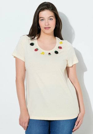 Kobieta o długich ciemnych włosach, ubrana w kremową koszulkę z krótkim rękawem, ozdobioną haftowanymi motywami owoców wokół dekoltu oraz niebieskie dżinsy.