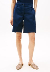 Granatowe bawełniane szorty o standardowym kroju, z kieszeniami z przodu i zapięciem na guzik. Gładka tekstura i długość do kolan. Noszone z beżowymi plecionymi klapkami.