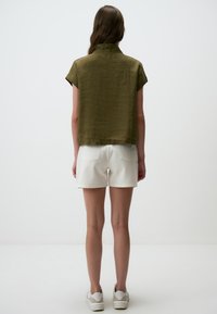 Camisetta corta a maniche corte verde oliva con colletto, realizzata in un tessuto testurizzato; abbinata a pantaloni corti bianchi e sneakers bianche.