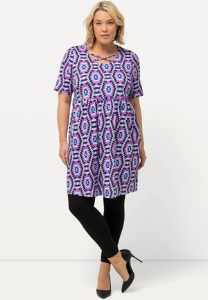 Lila- und pinkfarbendes, gemustertes Kleid mit kurzen Ärmeln, einem gefältelten Bund und einem kreuzförmigen Ausschnitt, kombiniert mit schwarzen Leggings und Absatzschuhen.