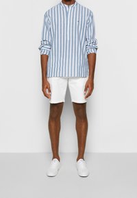 Camicia a maniche lunghe a righe blu chiaro e bianco con colletto alla mandarino, abbinata a pantaloni corti bianchi e sneakers bianche.
