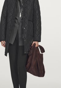 Giacca nera trapuntata con colletto alto, camicia grigia sotto e pantaloni neri larghi. Tiene in mano una borsa in suede marrone chiaro con manici.