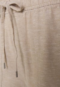 Ljust beige linne med en mjuk textur, utrustad med dragsko i midjan med metallspetsar och subtila vertikala mönster.