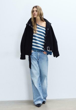 Žena má na sebe nadrozmernú čiernu bundu cez modrý a biely pruhovaný sveter so spustenými ramenami, voľné svetlomodré džínsy a čierne špicaté topánky.