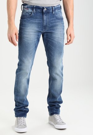 Homme portant un jean bleu délavé associé à des baskets gris clair, debout sur un fond blanc uni.