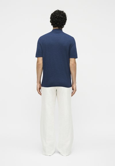 Άνδρας με σγουρά σκούρα μαλλιά, ντυμένος με μπλε σκούρο κοντομάνικο πόλο και άσπρο παντελόνι φαρδύ, στέκεται με την πλάτη γυρισμένη.