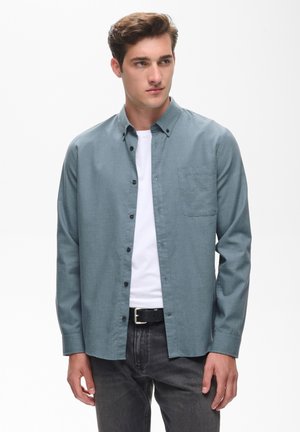 Junger Mann mit braunen Haaren, der ein blaues Hemd über einem weißen T-Shirt und schwarzen Hosen trägt und leicht zur Seite schaut.