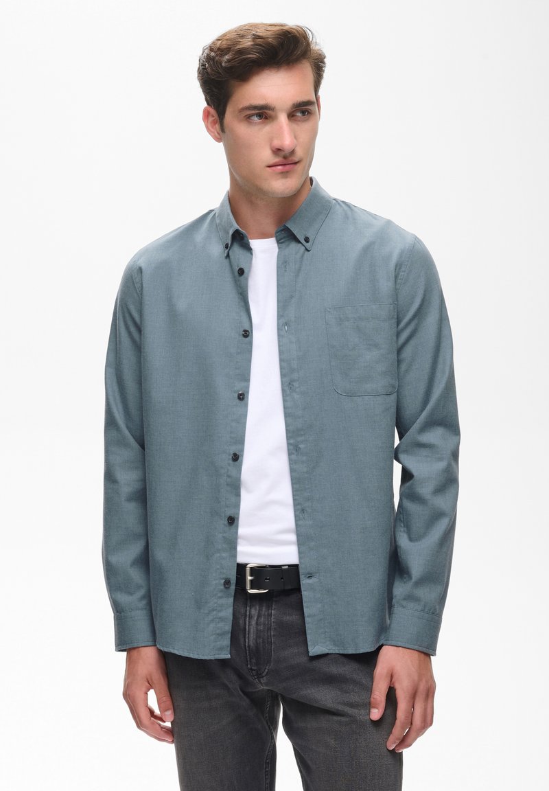 Jeune homme aux cheveux bruns portant une chemise bleue boutonnée par-dessus un t-shirt blanc et un pantalon noir, regardant légèrement sur le côté.