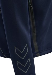 Veste de sport bleu marine à manches longues. Elle possède une poche zippée et des motifs en chevron gris sur les manches pour plus de détails.