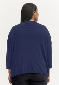 Sötétkék, oversized kötött pulóver háromnegyedes ujjal, lekerekített aljjal és oldalsó hasítékokkal a mozgás könnyebbsége érdekében. Simátokú textúra, lezser dizájn.