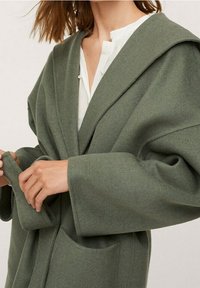 Woman adjusting the belt of an oversized olive green wool coat over a white blouse with pleated details.