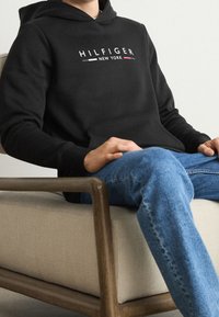 Svart hoodie med en vit "HILFIGER NEW YORK" logotyp, ficka framtill, ribbade muddar, bärs med ljusblå denimjeans, sitter på en beige stol.