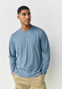 Long-sleeve shirt in soft blue fabric with round neckline. Features a small logo on the chest and a relaxed fit, paired with beige pants.