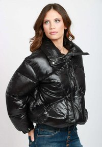 Giacca a vento nera con finitura lucida, caratterizzata da un colletto alto, cuciture in evidenza e chiusure a scatto. Indossata sopra jeans di denim.