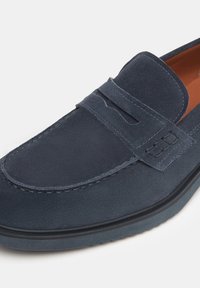 Marineblaue Wildleder-Loafer mit glatter Textur, versehen mit einem gesteppten Riemen und dezenten Akzenten, auf einer Gummisohle mit leichtem Absatz.