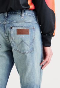 Šviesiai mėlyni Wrangler džinsai ant žmogaus, vilkinčio juodą ir oranžinę ilgomis rankovėmis marškinių, matomas užpakalinis kišenė ir ranka ilsisi šalia.