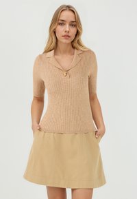 Haut beige à manches courtes et côtelé avec un col en forme de notch et un bouton en forme de lion doré, associé à une jupe A-line beige clair.