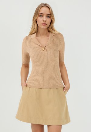 Haut beige à manches courtes et côtelé avec un col en forme de notch et un bouton en forme de lion doré, associé à une jupe A-line beige clair.