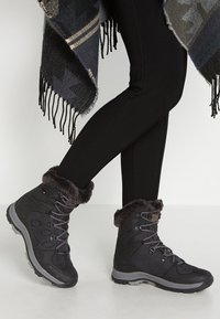 Black winter boots feature a faux fur cuff, textured fabric, and lace-up design. Soles are rugged for traction. Worn with black leggings.