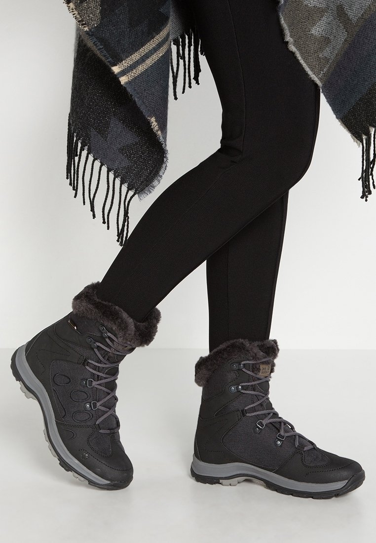 Black winter boots feature a faux fur cuff, textured fabric, and lace-up design. Soles are rugged for traction. Worn with black leggings.