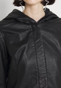 Veste en cuir noir avec capuche, présentant une surface texturée et des coutures verticales sur le devant. Aucune fermeture éclair ou ornement visible.