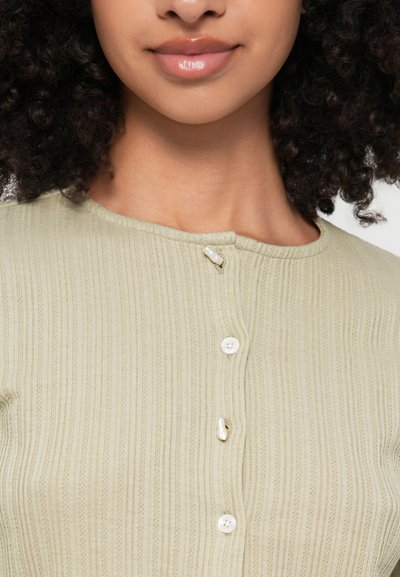 Ανοιχτό πράσινο ριπ πλεκτό γιλέκο με στρογγυλή λαιμόκοψη και τέσσερις κουμπιά, συμπεριλαμβανομένου ενός διακοσμητικού κουμπιού από αντίθετο υλικό.