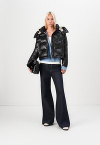 Schwarze Pufferjacke mit cropped Design und glänzender Textur, kombiniert mit einem blauen Strickjäckchen, dunklen, ausgestellten Jeans und einer schwarzen Clutch.