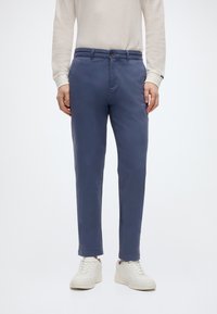 Μπλε παντελόνια από βαμβακερό chino με στενή εφαρμογή, δύο μπροστινές τσέπες, μία πίσω τσέπη και κλείσιμο με κουμπιά μπροστά. Φορεμένα με λευκά αθλητικά παπούτσια.