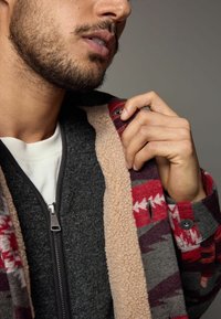Vêtements d'extérieur doublés en polaire avec un corps en laine grise, des manches à motifs rouges et une fermeture éclair. Comprend une couche intérieure crème pour une chaleur supplémentaire.