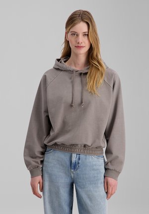 Frau mit langen blonden Haaren, die einen grauen Kapuzenpullover und hellblaue Jeans trägt, steht vor einem einfarbigen grauen Hintergrund.