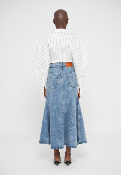 Persona con testa rasata che indossa una blusa bianca a righe verticali con maniche ampie e una lunga gonna di denim sbiadita, in piedi di spalle.