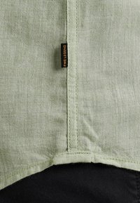 Lichtgroene stof met een textuurafwerking, voorzien van een naad en een klein zwart label met de tekst "PME Legend," met een zwarte rand eronder.