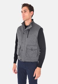 Gilet in misto lana grigio con colletto alto, dotato di due tasche anteriori con bottone e una tasca sul petto con zip, abbinato a una camicia nera a maniche lunghe.