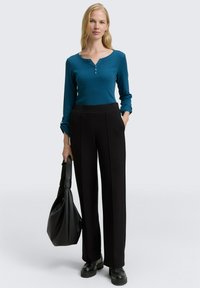 Vrouw in een teal lange mouw top, zwarte hoge taille broek, zwarte schoenen, met een zwarte handtas, staand tegen een effen witte achtergrond.