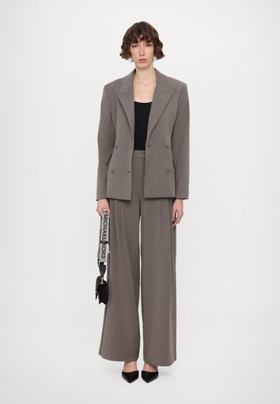 Grauer, doppelt geknöpfter Blazer kombiniert mit weit geschnittenen grauen Hosen. Das Model hält eine kleine schwarze Tasche mit einem Logostreifen und trägt schwarze spitze Schuhe.