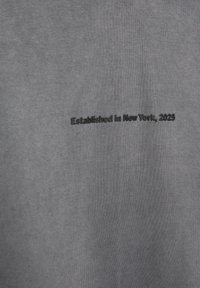 Szary materiał o miękkiej teksturze z haftowanym napisem "Założono w Nowym Jorku, 2025" w ciemnym kolorze.