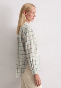 Lekka, w kratę koszula w kolorze beżowym i niebieskim, z kołnierzykiem na guziki i długimi rękawami. Boczny rozpór w dolnej części podkreśla wentylację.