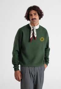 Zielony sweter z dzianiny z prążkowanymi mankietami, z żółtym haftem na piersi, zestawiony z wzorzystym jedwabnym szalem i paskami.