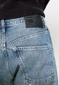 Dżinsy denim w jasnoniebieskim kolorze z efektem wyblakłego wykończenia, z czarną skórzaną naszywką marki na talii oraz dwoma tylnymi kieszeniami z ozdobnymi przeszyciami.