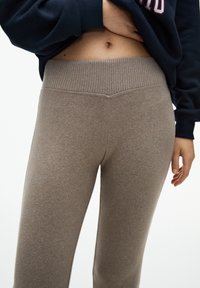 Close-up van een persoon die beige, hooggeregde ribgebreide broek en een donkere sweatshirt draagt, terwijl de hand het shirt optilt om de buik te onthullen.