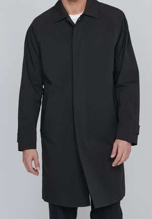 Homme portant un long manteau noir par-dessus une chemise blanche, debout devant un fond gris uni.