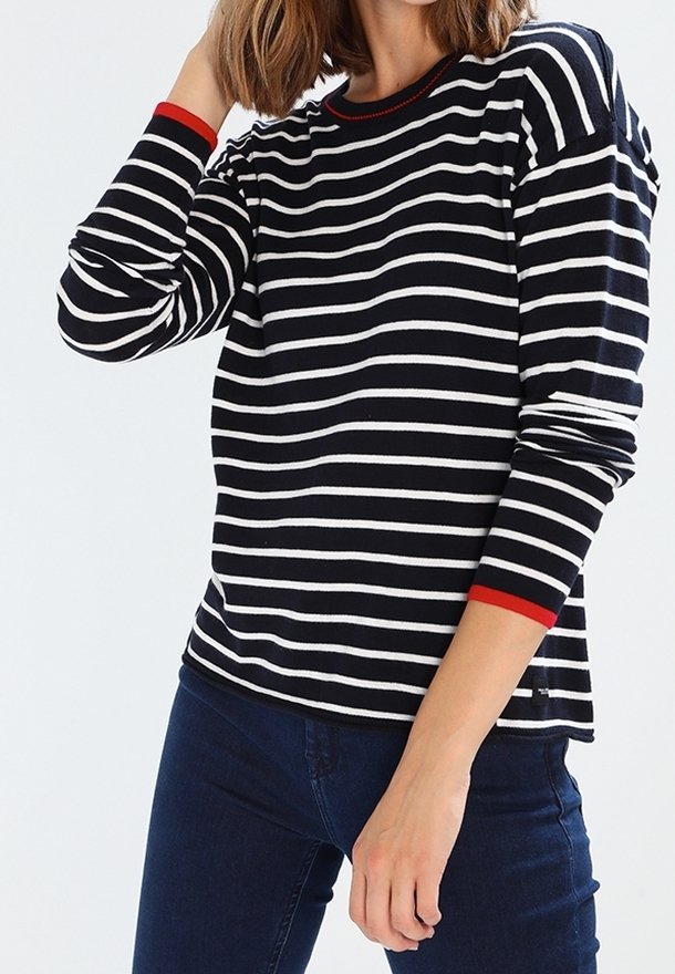 Femme portant une chemise à manches longues rayée horizontalement en noir et blanc avec un bord rouge aux poignets et au col, associée à un jean bleu foncé.