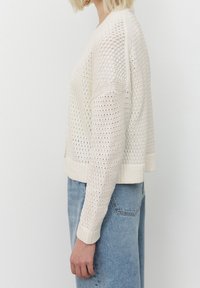 Cardigan leggero di colore crema, lavorato a maglia con una trama aperta, design corto, con polsini e bordo a costine, indossato sopra jeans azzurri chiari.